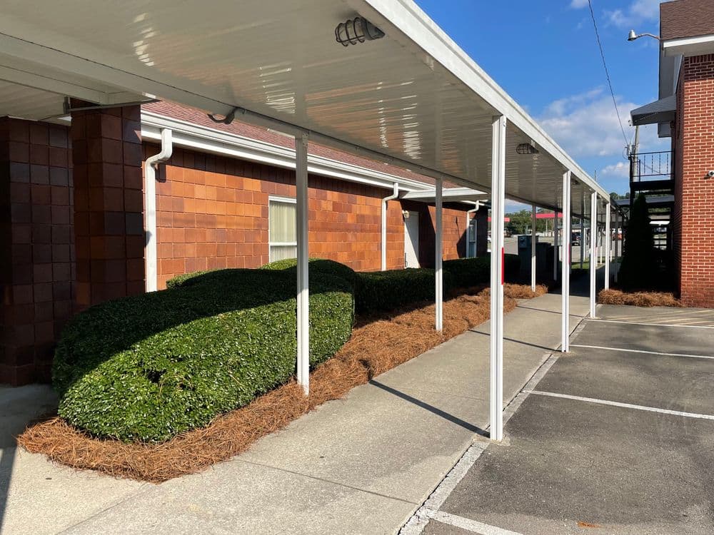 Walkway with covered awning alongside neatly trimmed bushes and fresh pine straw mulch.