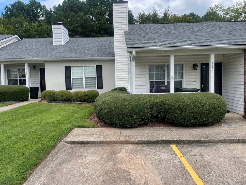 Single-story duplex with neatly trimmed bushes and parking spaces, surrounded by green grass.