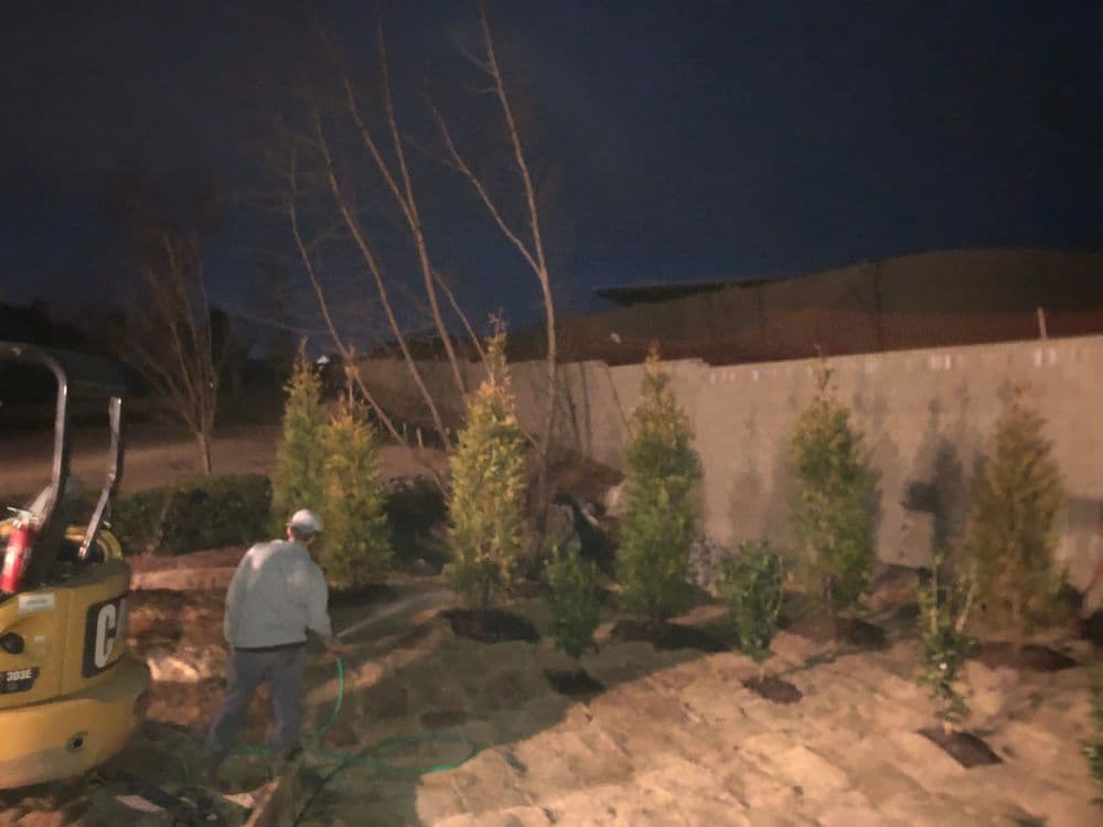 Person watering newly planted trees in a landscaped area at night. Heavy machinery nearby.