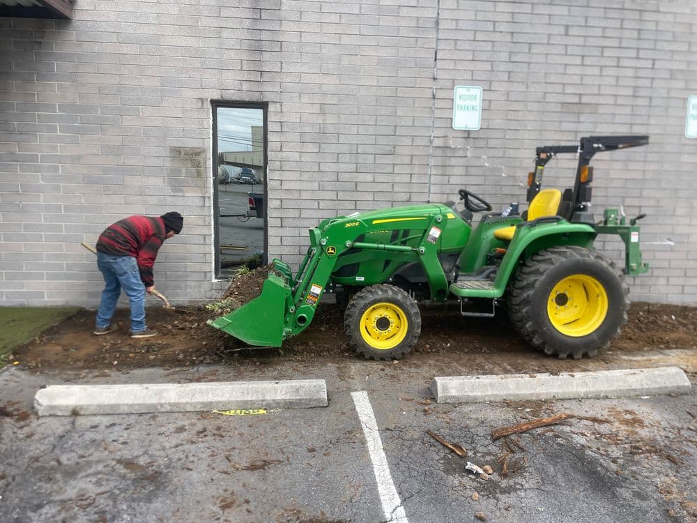 Man operating a green John Deere tractor near a building, clearing debris from the ground.