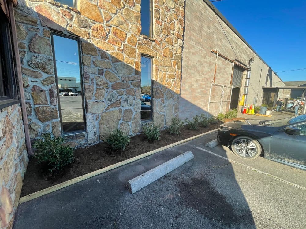 Landscaped area with greenery next to a stone building and parked car.