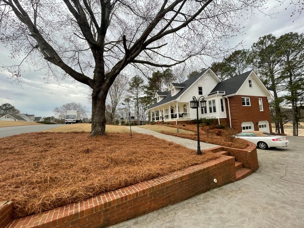 Elegant brick house with a large front porch, tree, and landscaped driveway in a suburban setting.