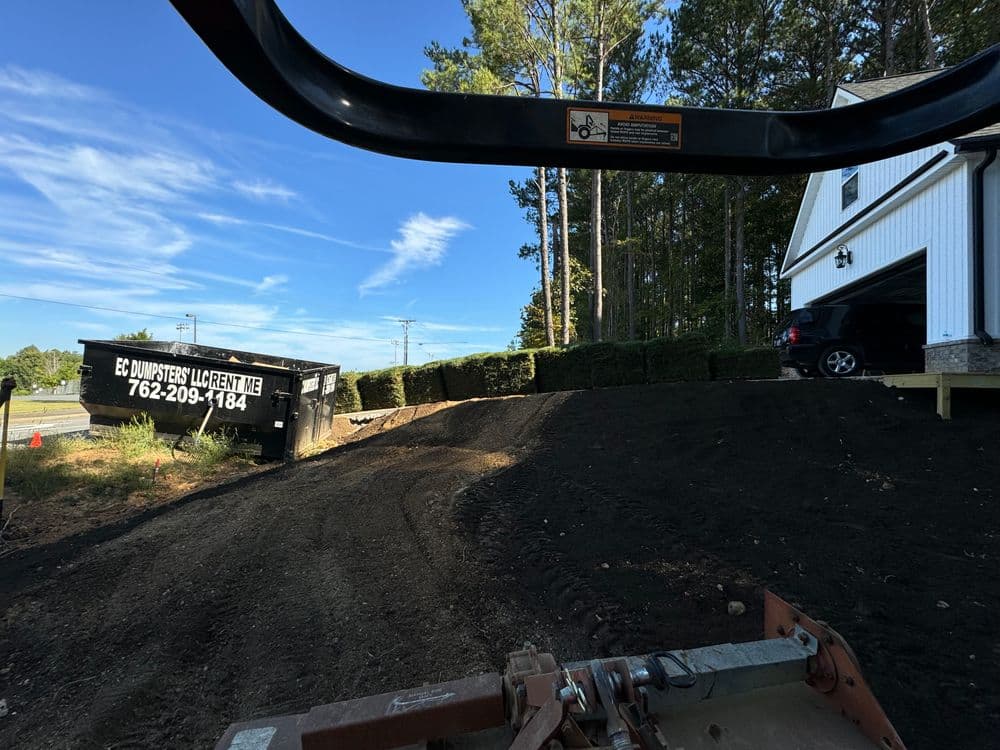 Construction scene with a view of rolled sod next to a house and dumpster, under a clear blue sky.