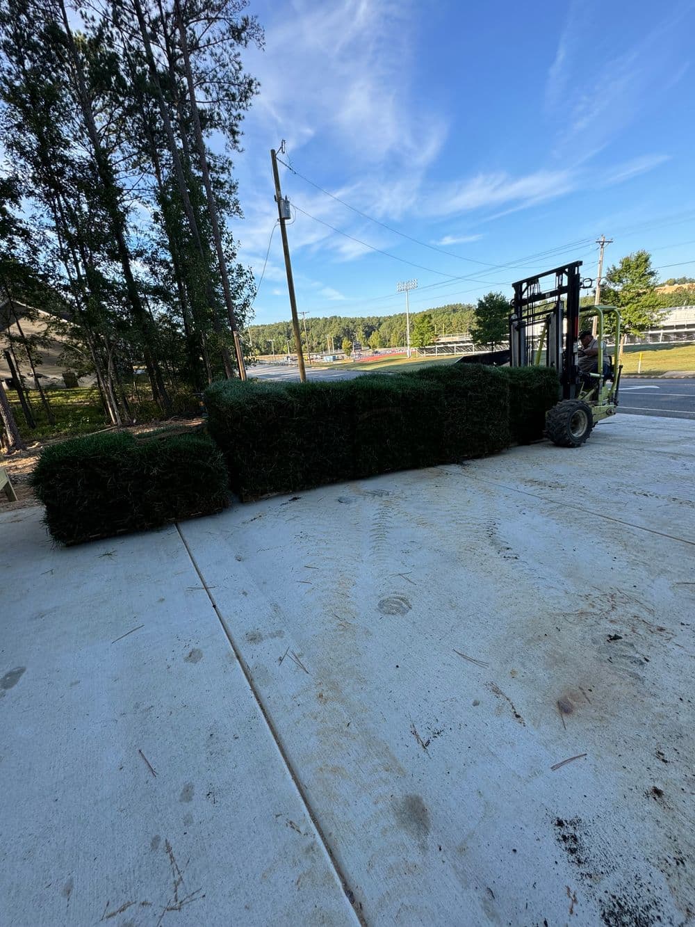 Forklift delivering hay bales on a sunny day near a sports field and wooded area.