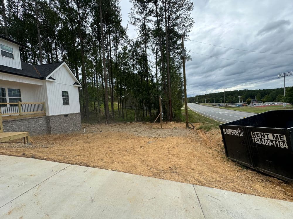 Newly cleared land next to a house, surrounded by trees and a construction dumpster nearby.