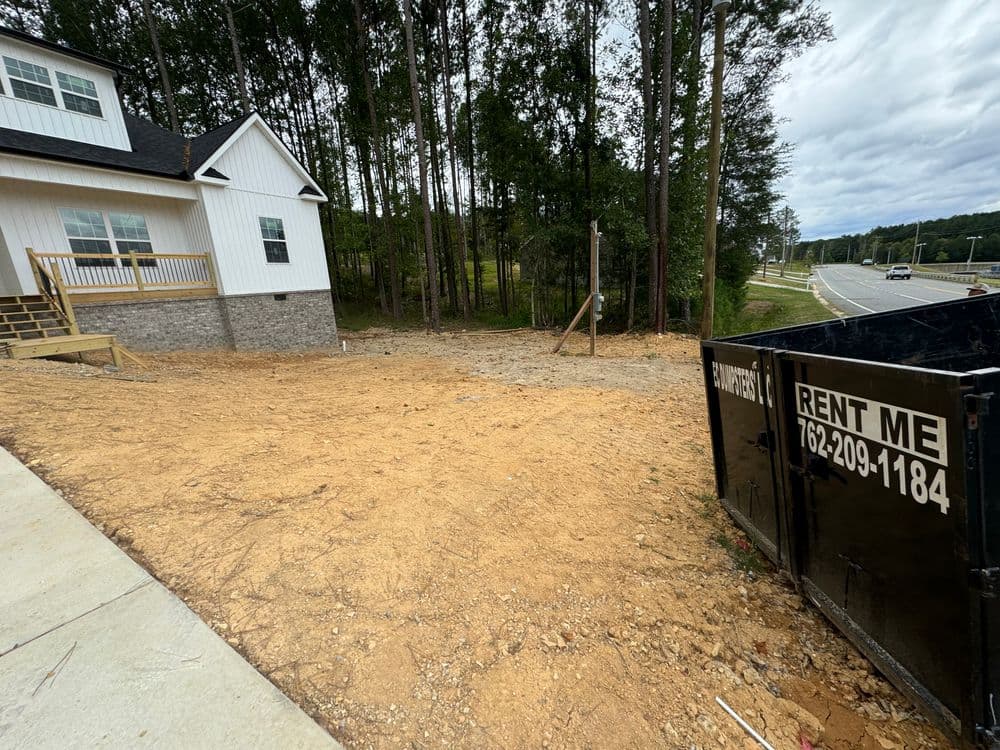 New construction site with dumpster for debris on a wooded road. Contact for rental services.