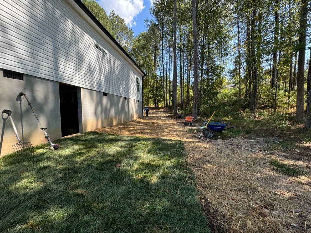 Lawn maintenance view beside a house, featuring a wheelbarrow and tools in a wooded area.
