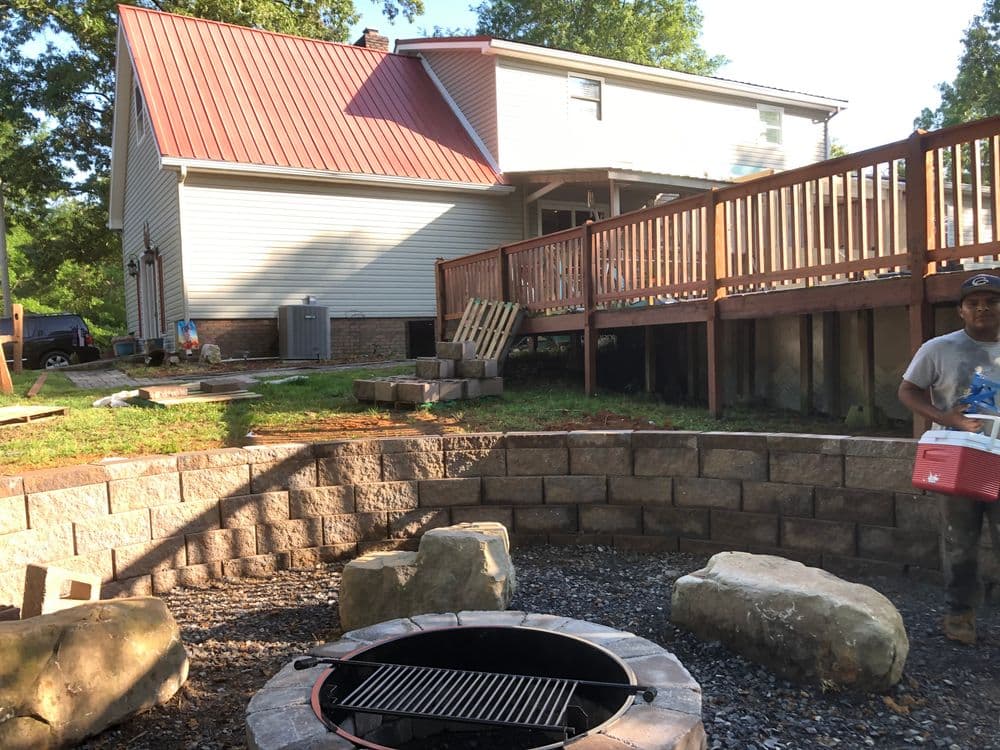 Backyard setting with a fire pit, stone seating, and a house with a deck in the background.