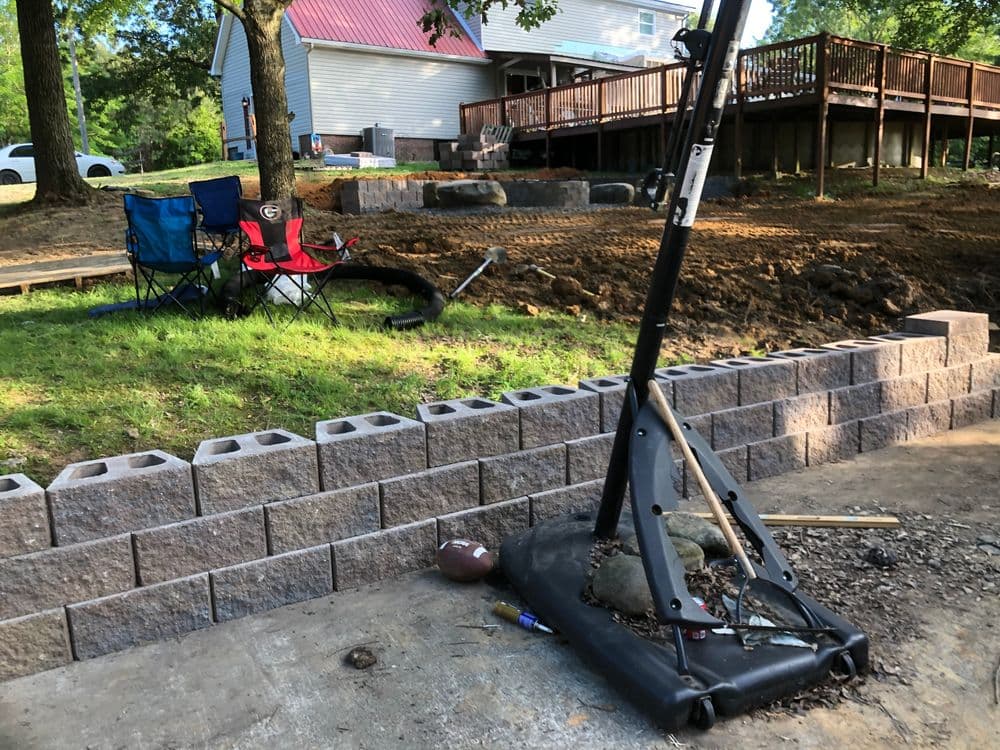 Basketball hoop on a driveway with stone wall, chairs, and yard construction in background.