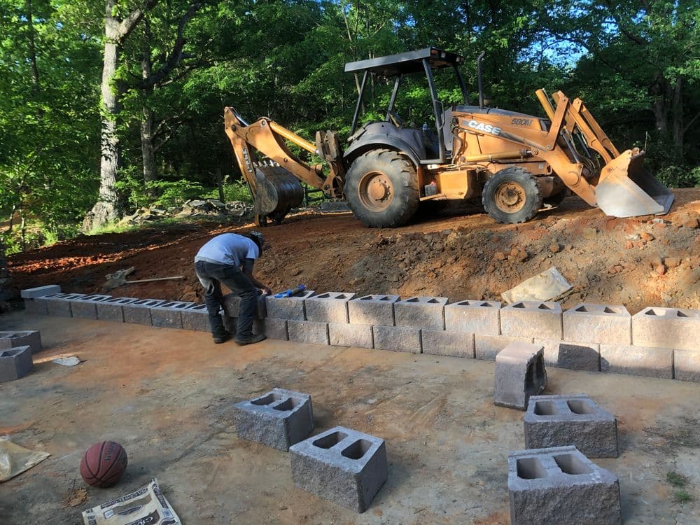 Construction worker laying cinder blocks with a CASE backhoe in a wooded area.