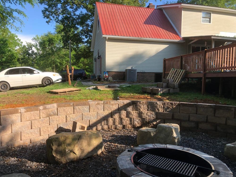 Backyard fire pit area with stone walls, car parked nearby, and a house with a red roof.